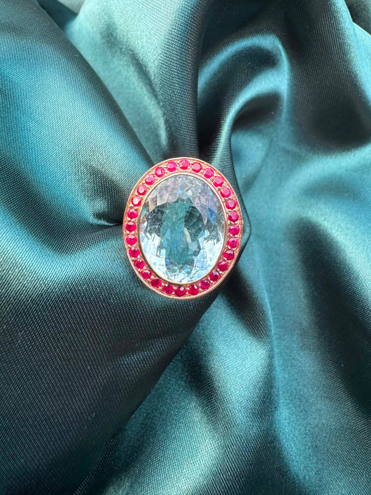Aquamarine and gemstone halo ring in 14ct rose gold on teal fabric, front view showing oval aquamarine and red gemstone halo