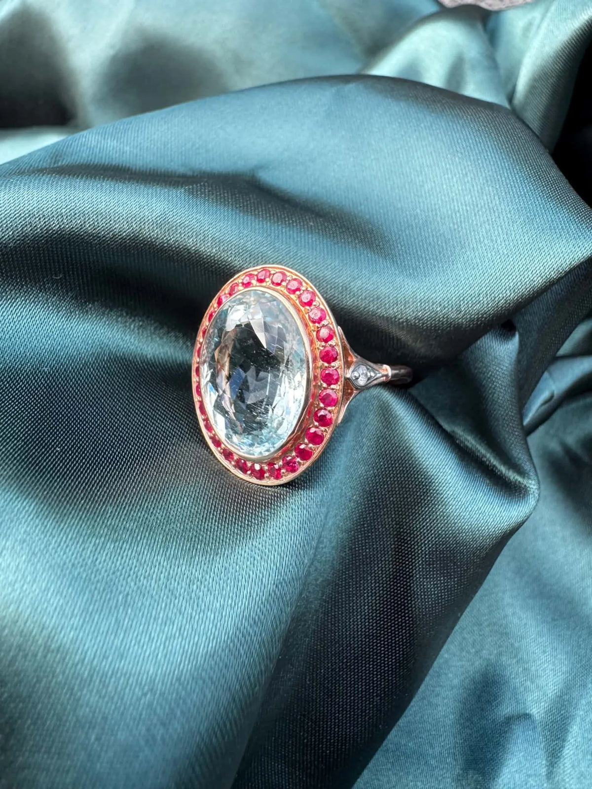 Aquamarine and gemstone halo ring in 14ct rose gold, angled top-side view showing oval aquamarine in red gemstone halo setting