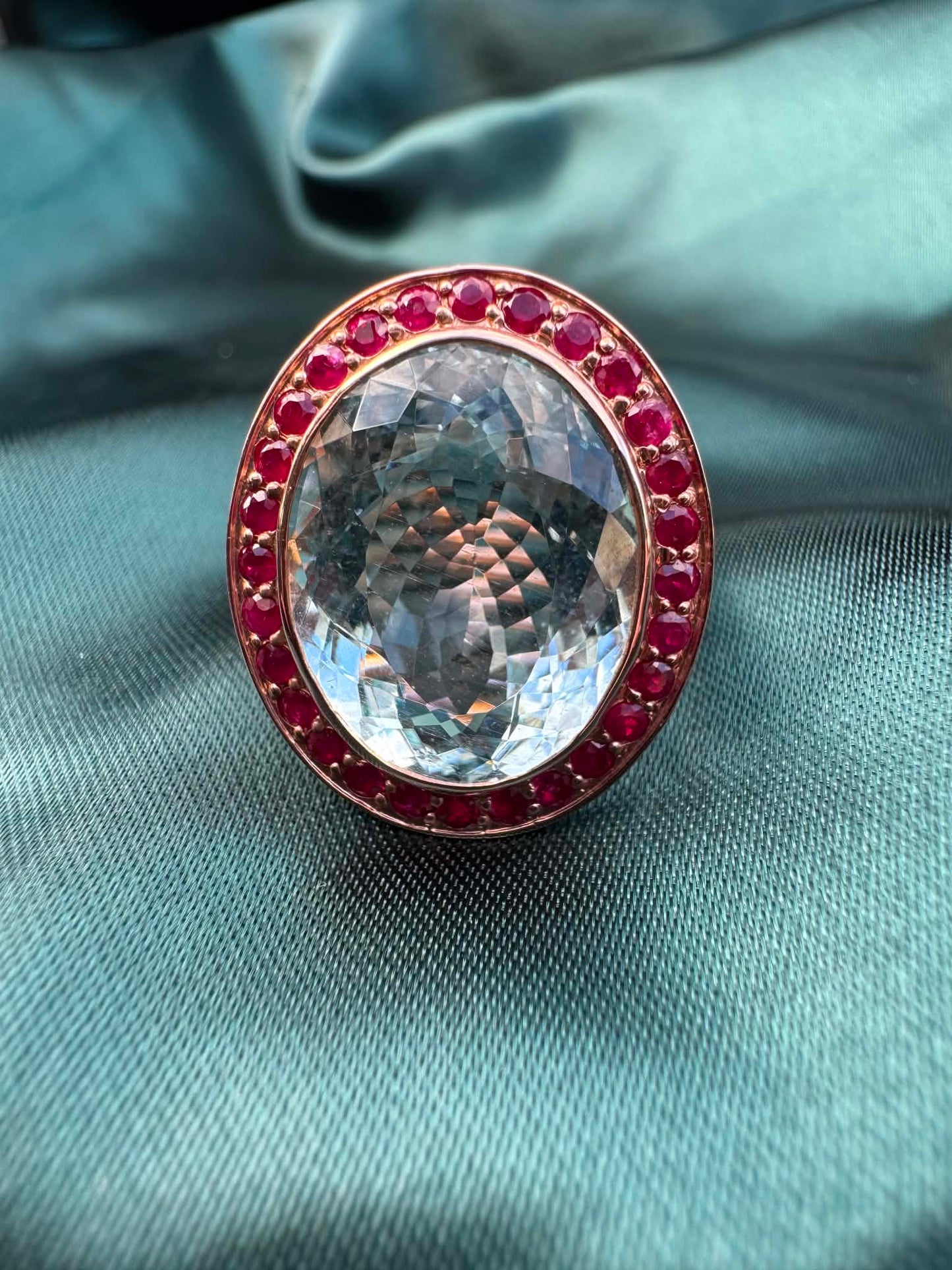 Aquamarine and gemstone halo ring in 14ct rose gold on teal fabric, close-up showing oval aquamarine and red gemstone halo